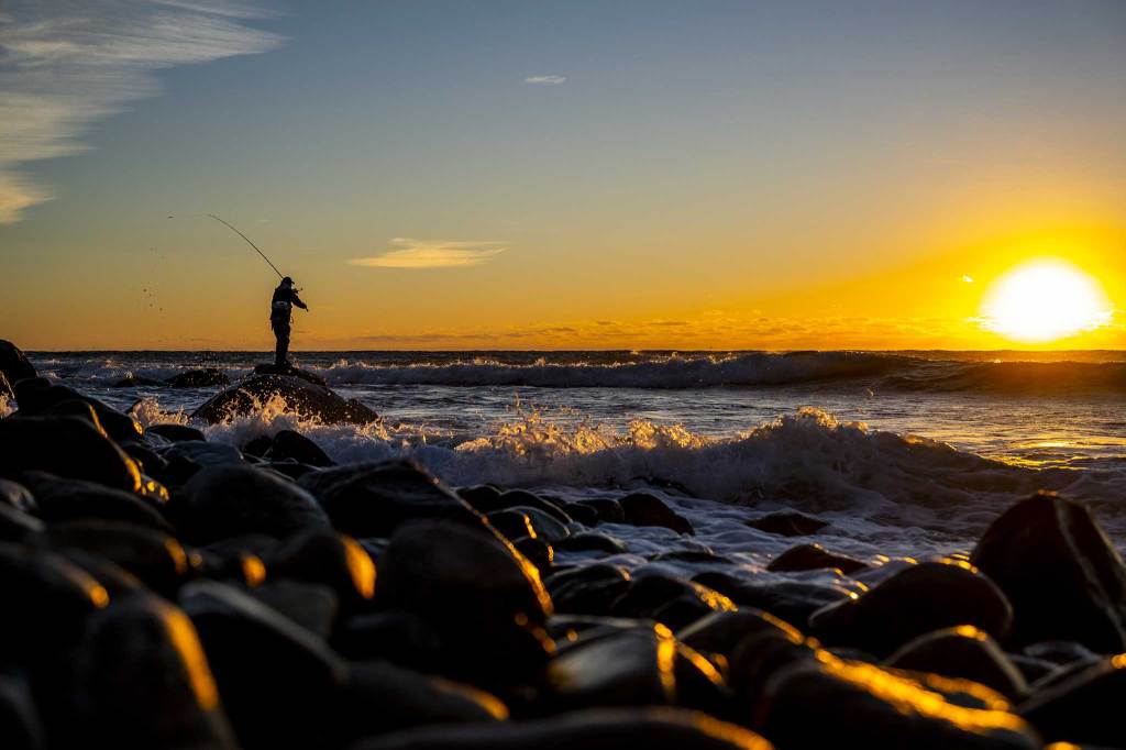 A fisherman on a rocky coast at sunrise casting a rod.