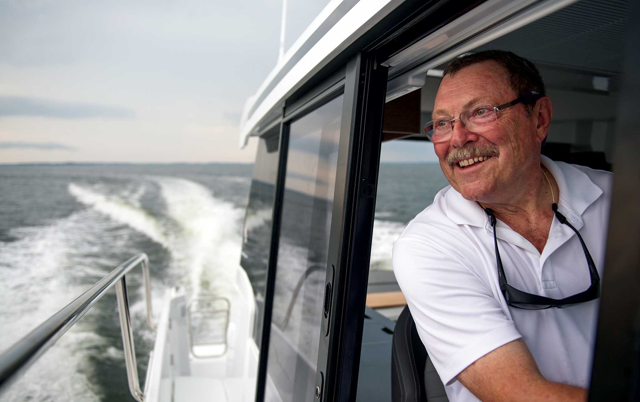 A smiling man looking out the side boat window as it leaves a wake.