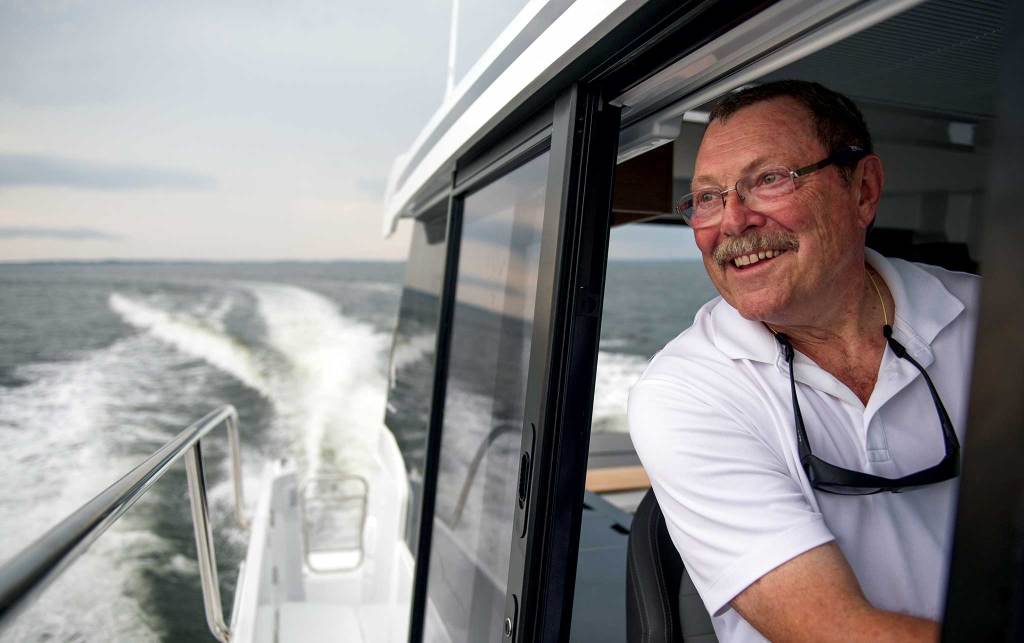 Jeanneau-Selects-PMY-9.2018-©-Jay-Fleming05-2048x A smiling man looking out the side boat window as it leaves a wake.